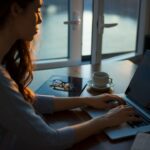 Person arbetar vid laptop med kaffe och glasögon vid ett fönster