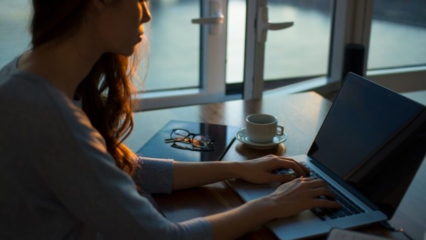 Person arbetar vid laptop med kaffe och glasögon vid ett fönster