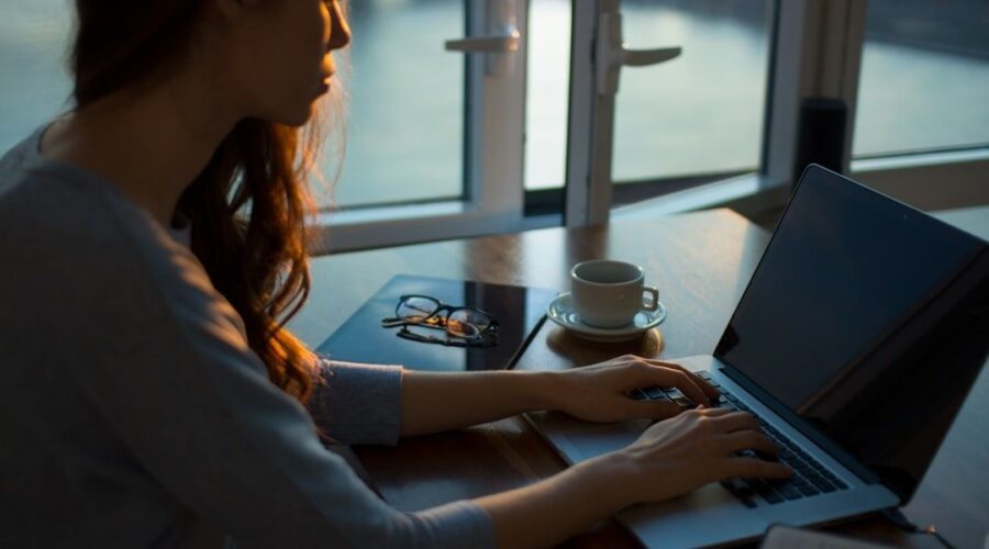 Person arbetar vid laptop med kaffe och glasögon vid ett fönster