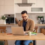 Man arbetar vid laptop i ett kök, med kaffekopp och anteckningsbok