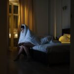 Vaken person sitter på sängkanten insvept i täcke under natten
