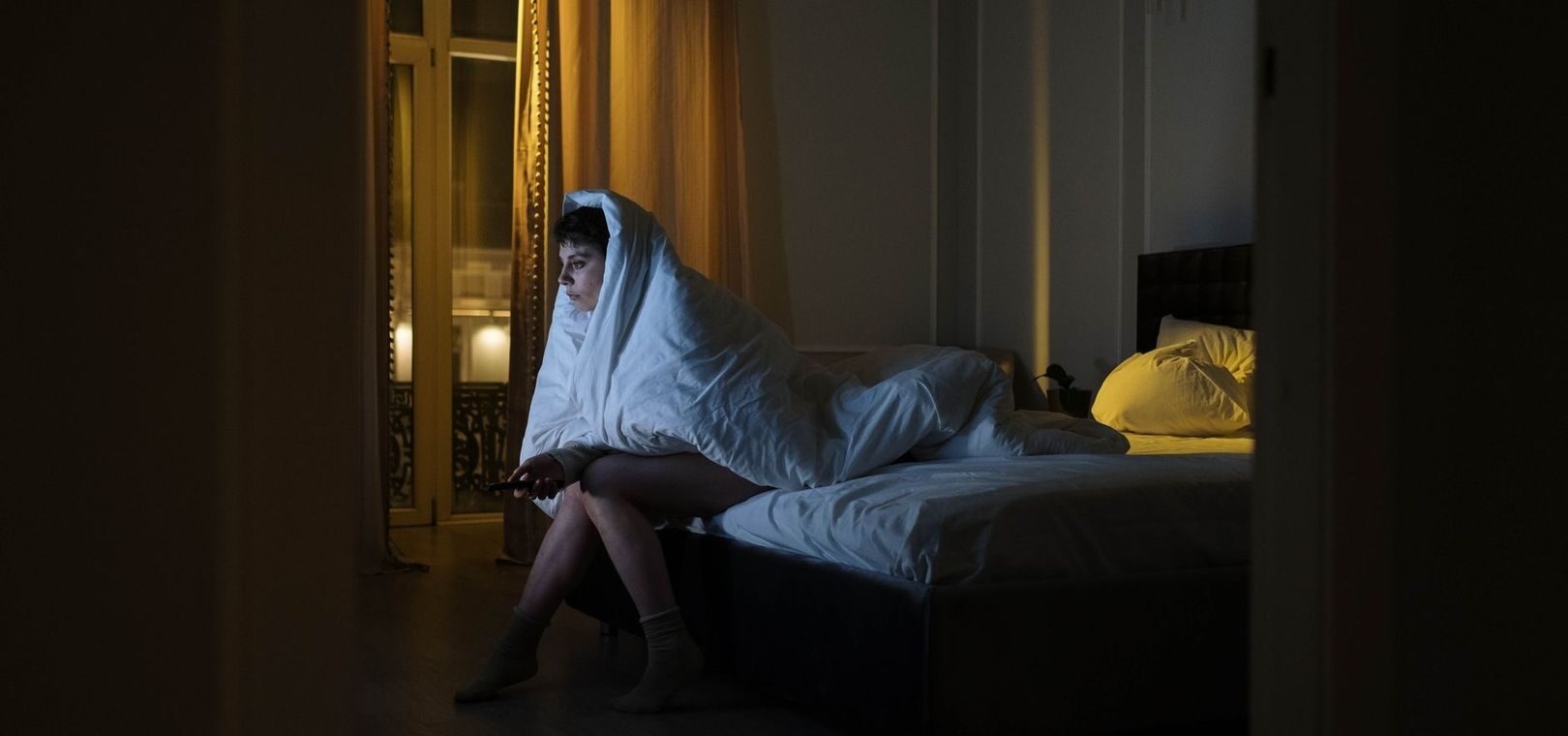 Vaken person sitter på sängkanten insvept i täcke under natten