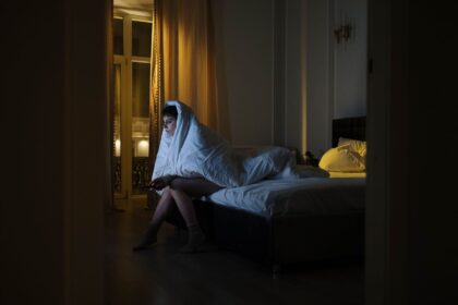 Vaken person sitter på sängkanten insvept i täcke under natten