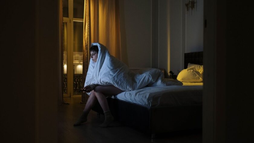 Vaken person sitter på sängkanten insvept i täcke under natten